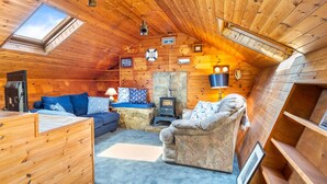 Un interior de cabaña acogedor con techo de madera, chimenea de piedra, un sofá, una silla y un televisor.