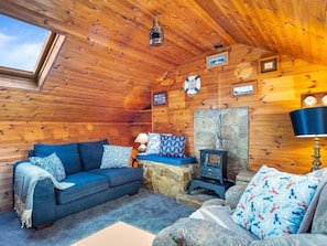 A cozy cabin interior with a wooden ceiling, a stone wall, a blue sofa, a table with a lamp, and a wood-burning stove.