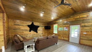 A log cabin interior with a brown sofa, a white coffee table, and a mounted animal hide on the wall. - The Bear Den ATV friendly (Mena)