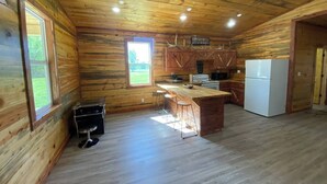 A log cabin kitchen with wooden walls, a central island with stools, a refrigerator, and a window overlooking a green landscape. - The Bear Den ATV friendly (Mena)