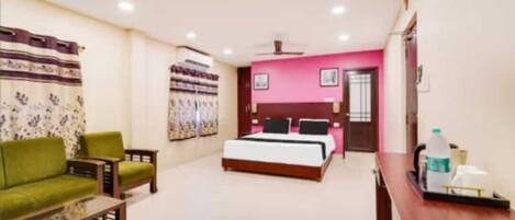 A hotel room with a bed, a green sofa, a wooden coffee table, a desk, and a pink accent wall.