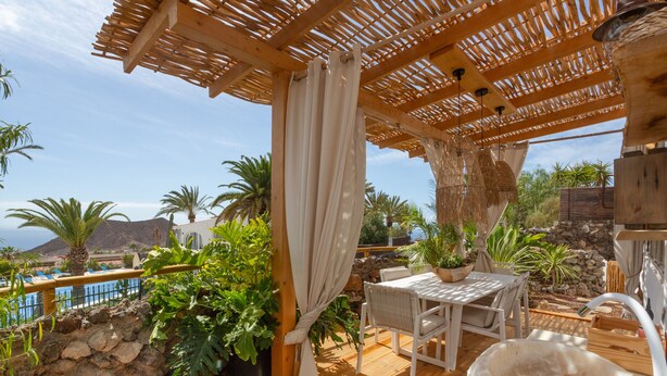 Una terraza de madera con un muro de piedra, una mesa con sillas, plantas en macetas y vista al mar.