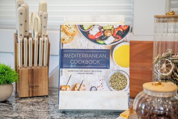 A Mediterranean cookbook on a kitchen counter with a knife block, a glass jar, and a potted plant.