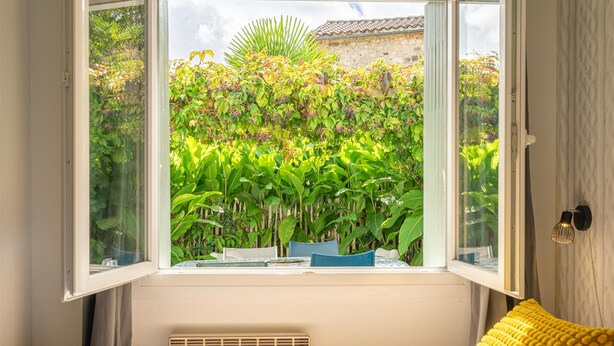 A bedroom with a window view of a lush garden, a bed with a yellow pillow, and a radiator. - Holiday Home 'Du Porche' with Private Terrace and Wi-Fi (Chaillevette)