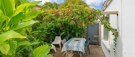 A patio with a table and chairs, surrounded by lush greenery and a white wall.
