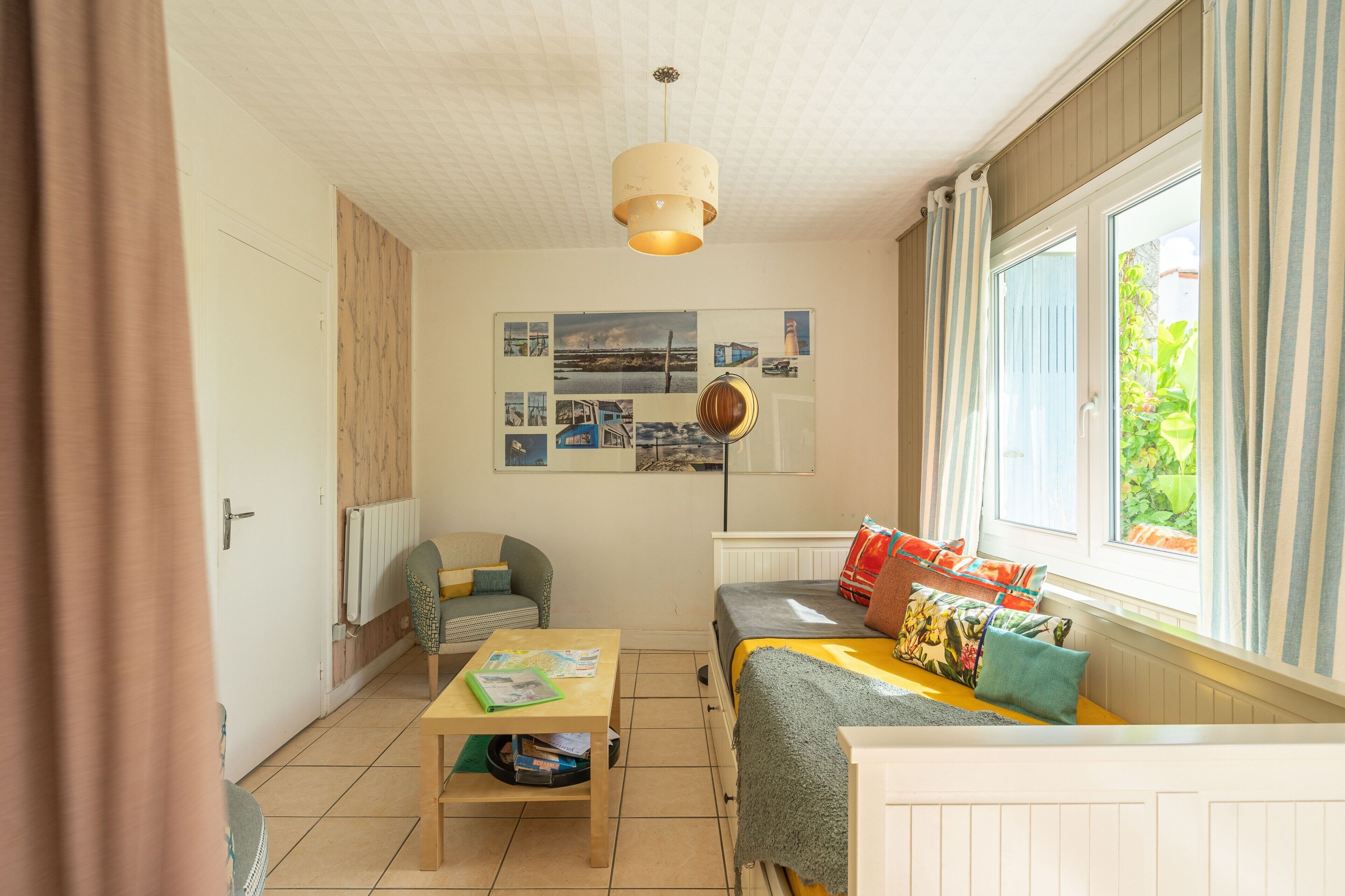 A room with a bed, a table, a chair, a radiator, a window with curtains, and a wall with pictures.