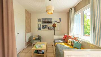 A room with a bed, a table, a chair, a radiator, a window with curtains, and a wall with pictures.