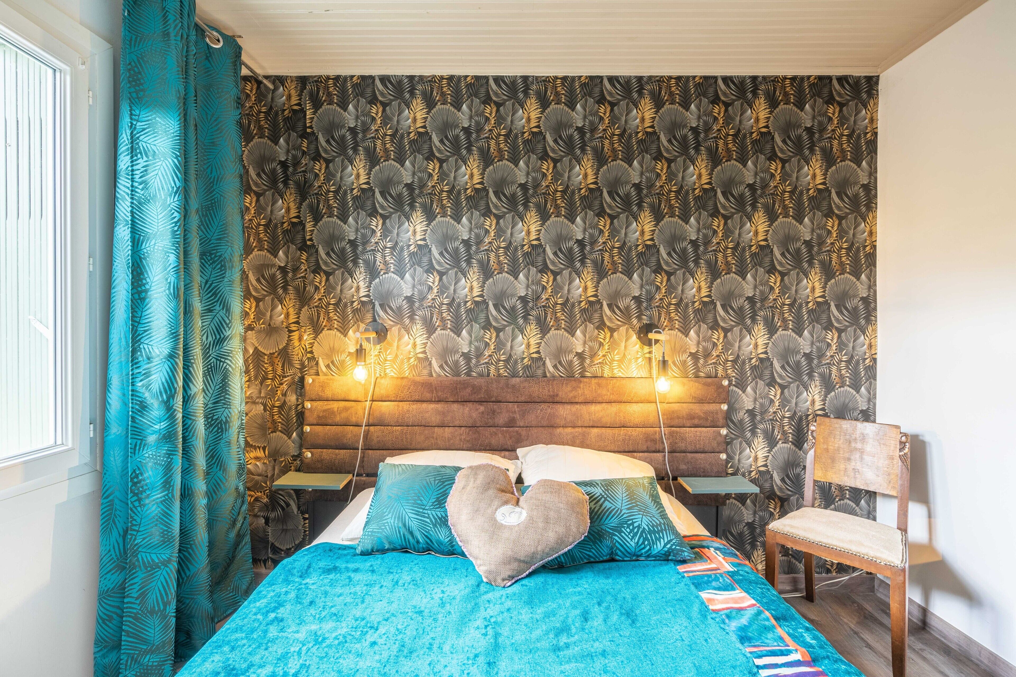 Ein Schlafzimmer mit gemustertem Wandpapier, einem hölzernen Kopfteil, einer türkisblauen Bettdecke und einem herzförmigen Kissen.