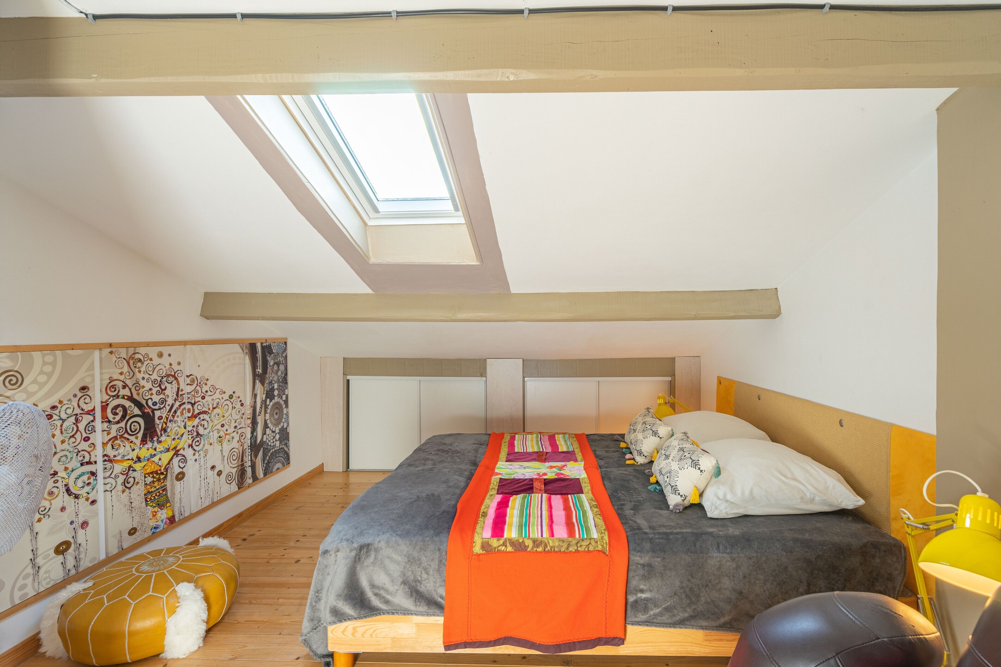 A bedroom with a skylight, a bed with a colorful blanket, a headboard, and a wall with a patterned tapestry.