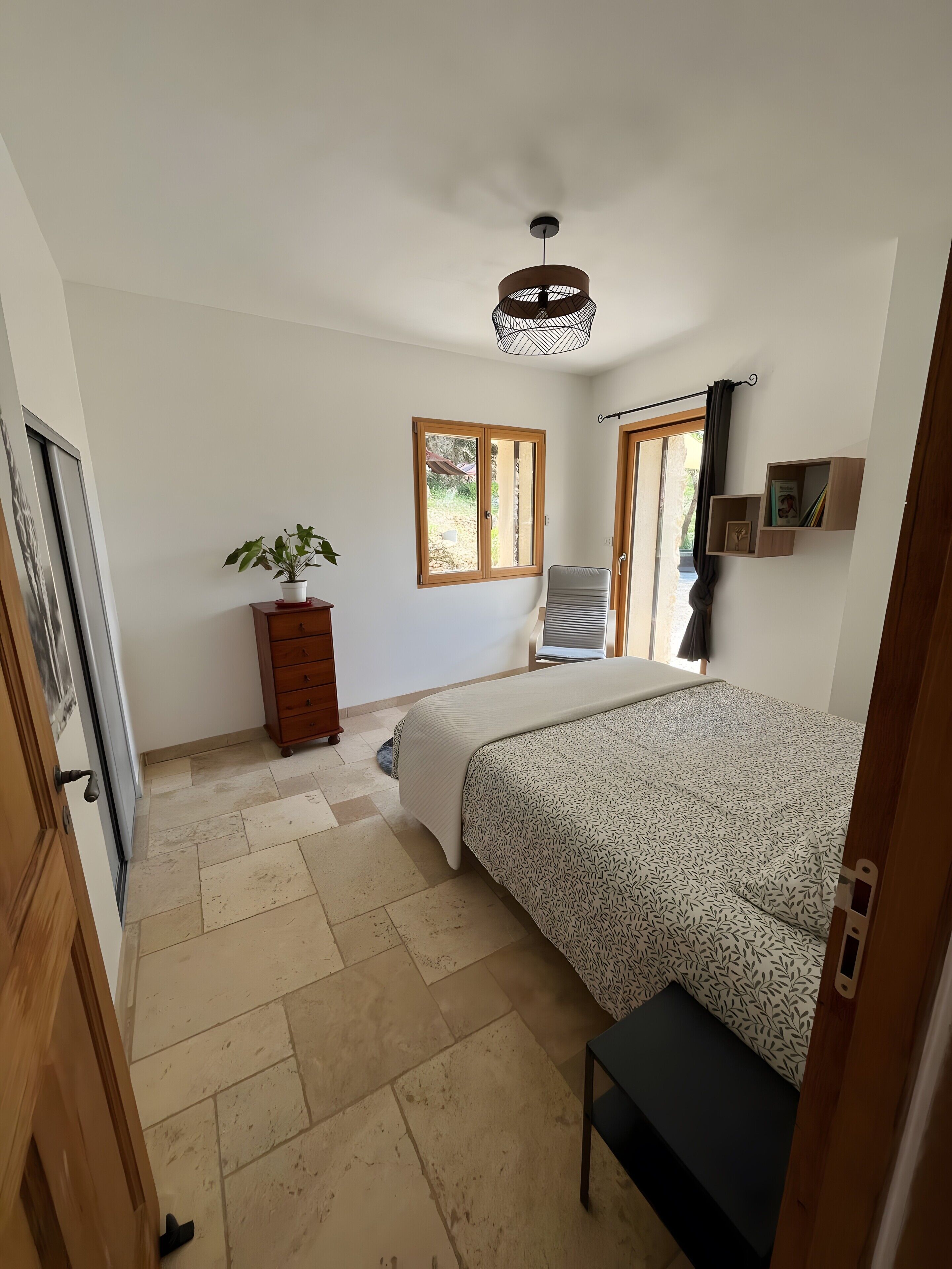 A bedroom with a bed, a chair, a dresser, a potted plant, a window, and a ceiling light fixture.