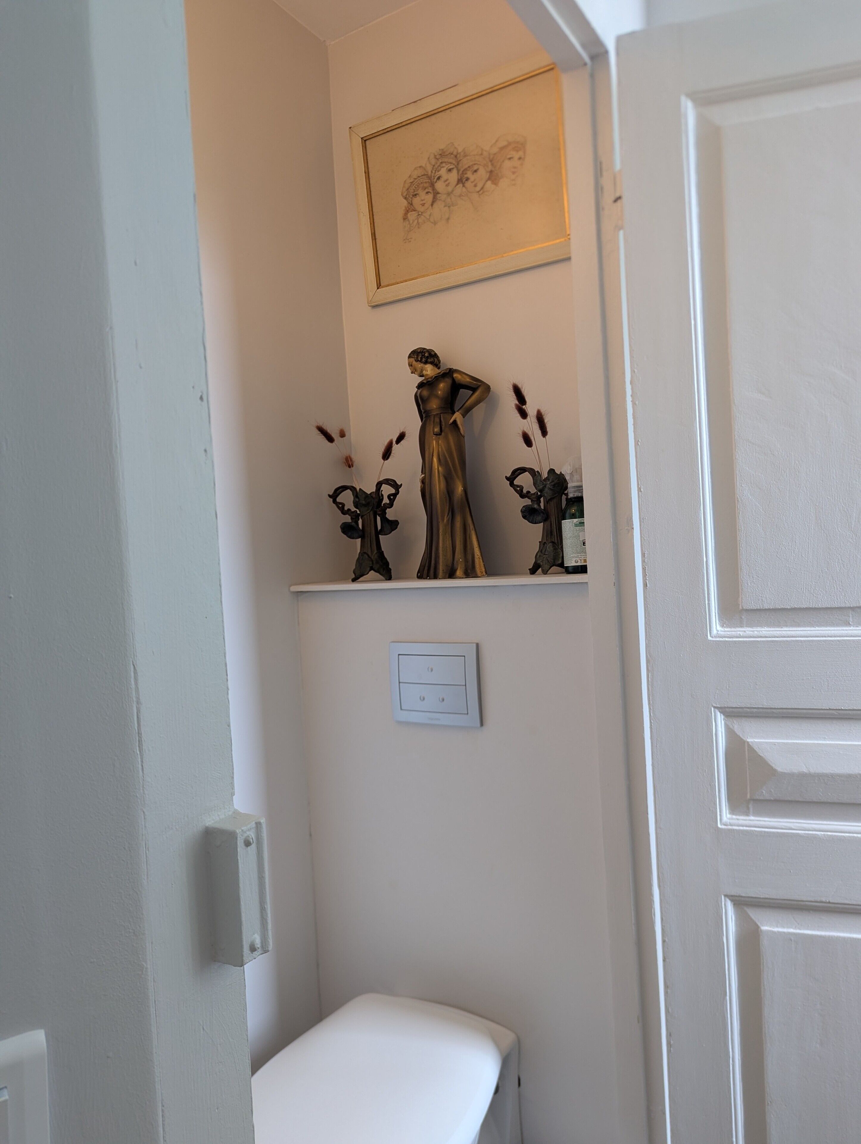 A bathroom with a white bench, a wall-mounted light switch, and a framed picture above a decorative statue.