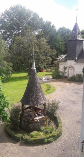 A gazebo with a conical roof, surrounded by a circular hedge, in a garden area with a church in the background. - Château De Chaunac - Room 1 (Naves)