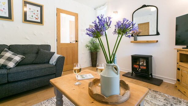Un salón acogedor con un sofá gris, una mesa de centro de madera con un jarrón de flores moradas, una chimenea y un televisor sobre un mueble de madera.