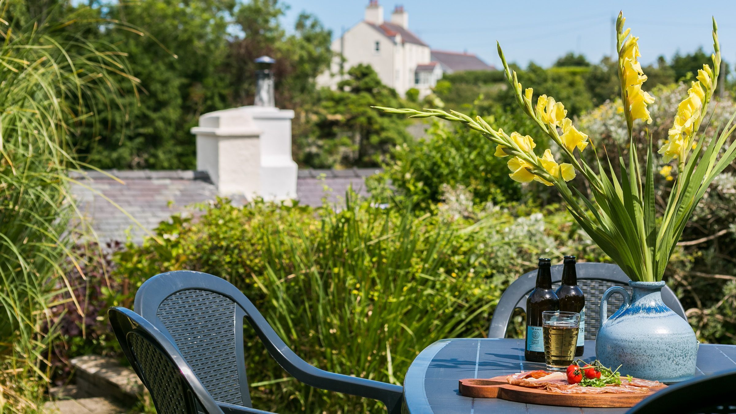 A table set outdoors with a bottle of wine, a glass, a vase with yellow flowers, and a plate with food.