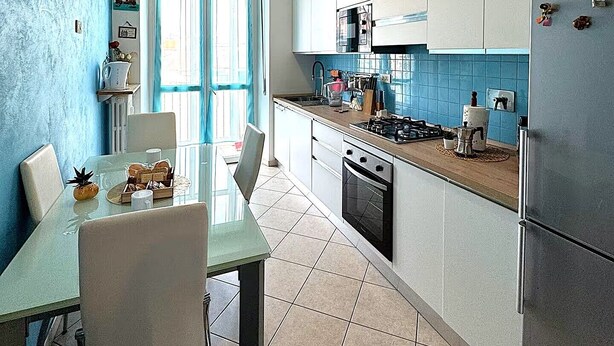 A modern kitchen with white cabinets, a gas stove, and a refrigerator. There is a dining table with chairs and a window with blue curtains.