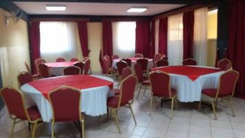 Un salón de conferencias con varias mesas y sillas, ventanas amplias con cortinas rojas y piso de baldosas.