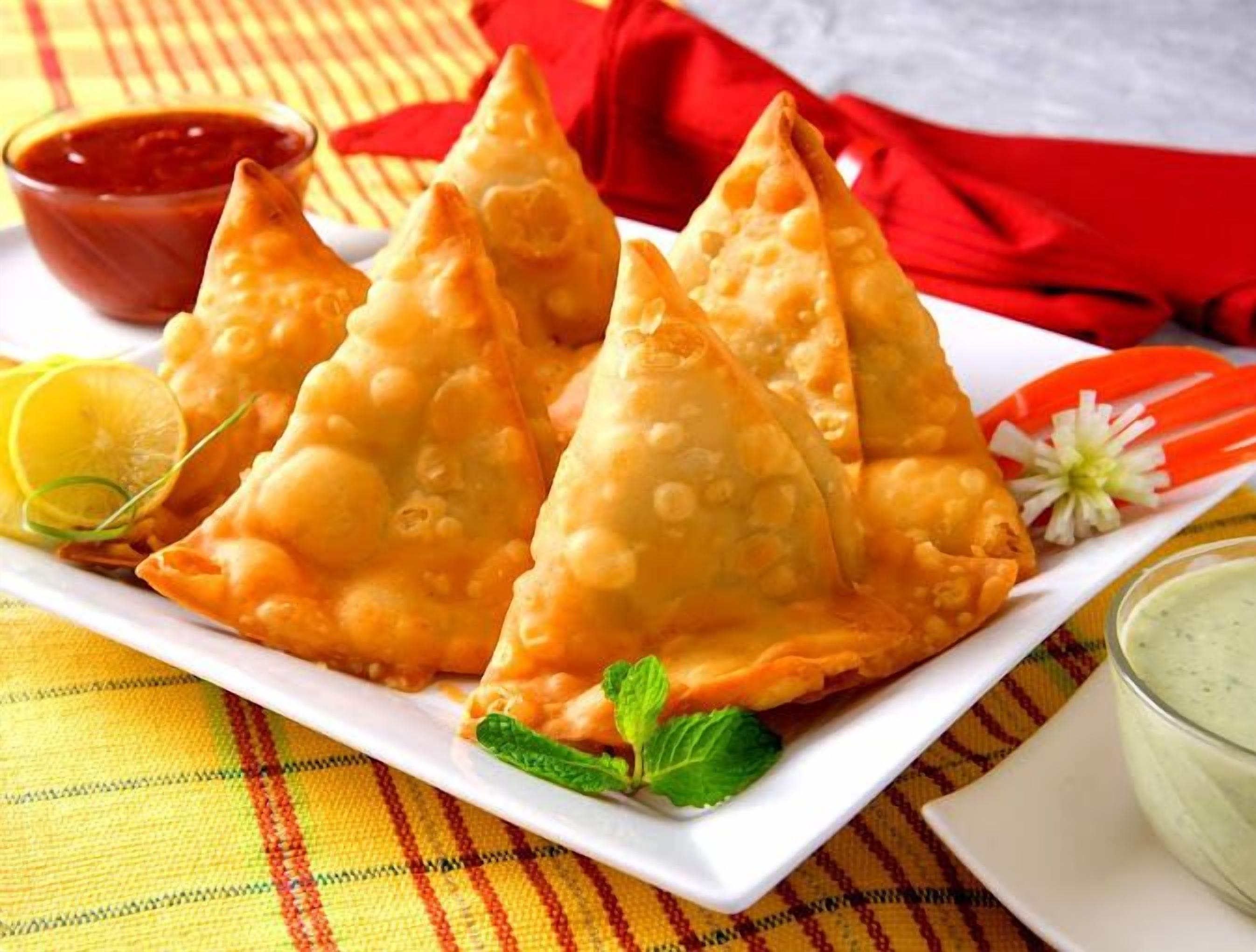 Un plato de samosas con gajos de limón y hojas de menta, acompañado de dos salsas para mojar.