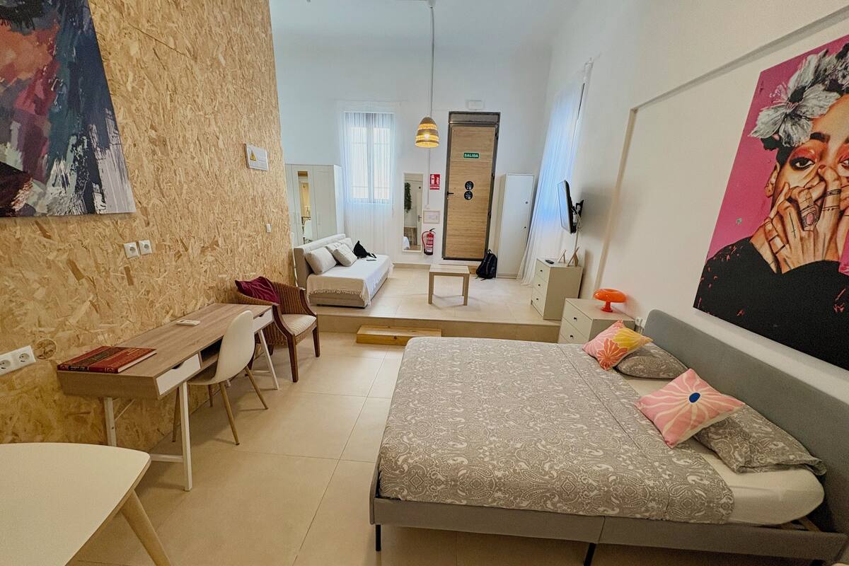 A modern bedroom with a bed, desk, chair, and a sofa. There is a painting on the wall and a door with a sign.