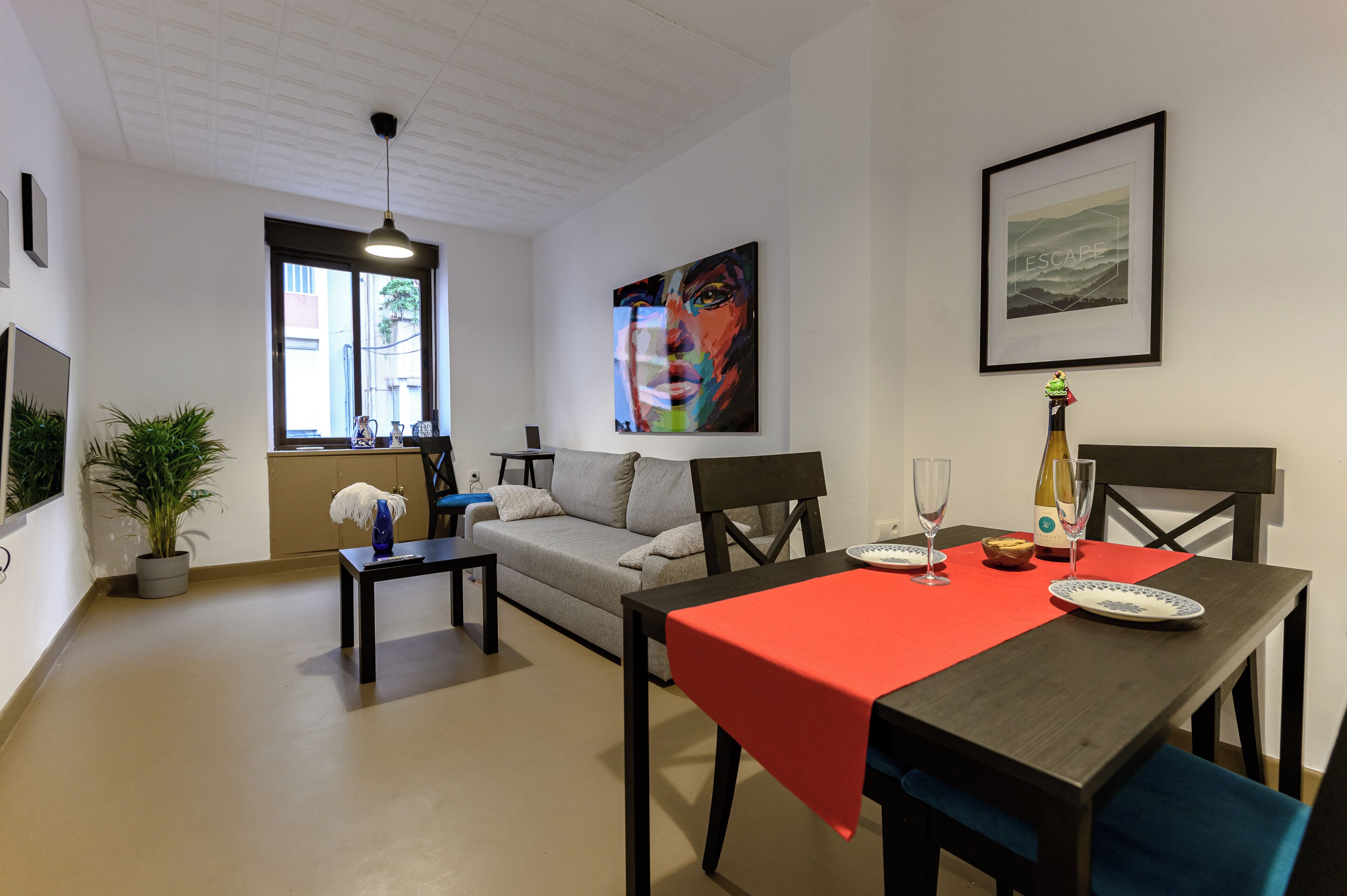 Un salon moderne avec un canapé, une table à manger recouverte d’une nappe rouge et une fenêtre donnant sur des immeubles.