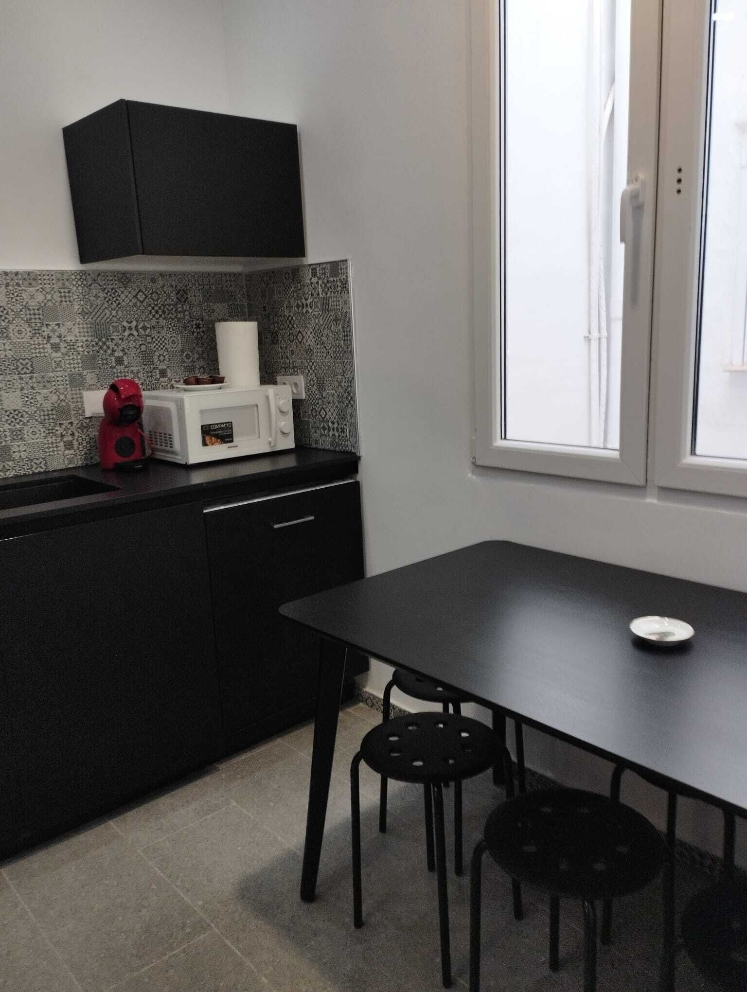 A compact kitchen with a microwave, a kettle, and a black cabinet. There is a dining table with two chairs and a window with blinds.
