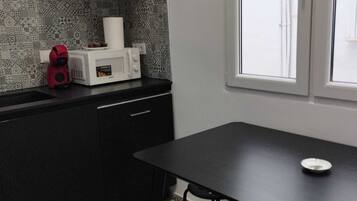 A compact kitchen with a microwave, a kettle, and a black cabinet. There is a dining table with two chairs and a window with blinds.