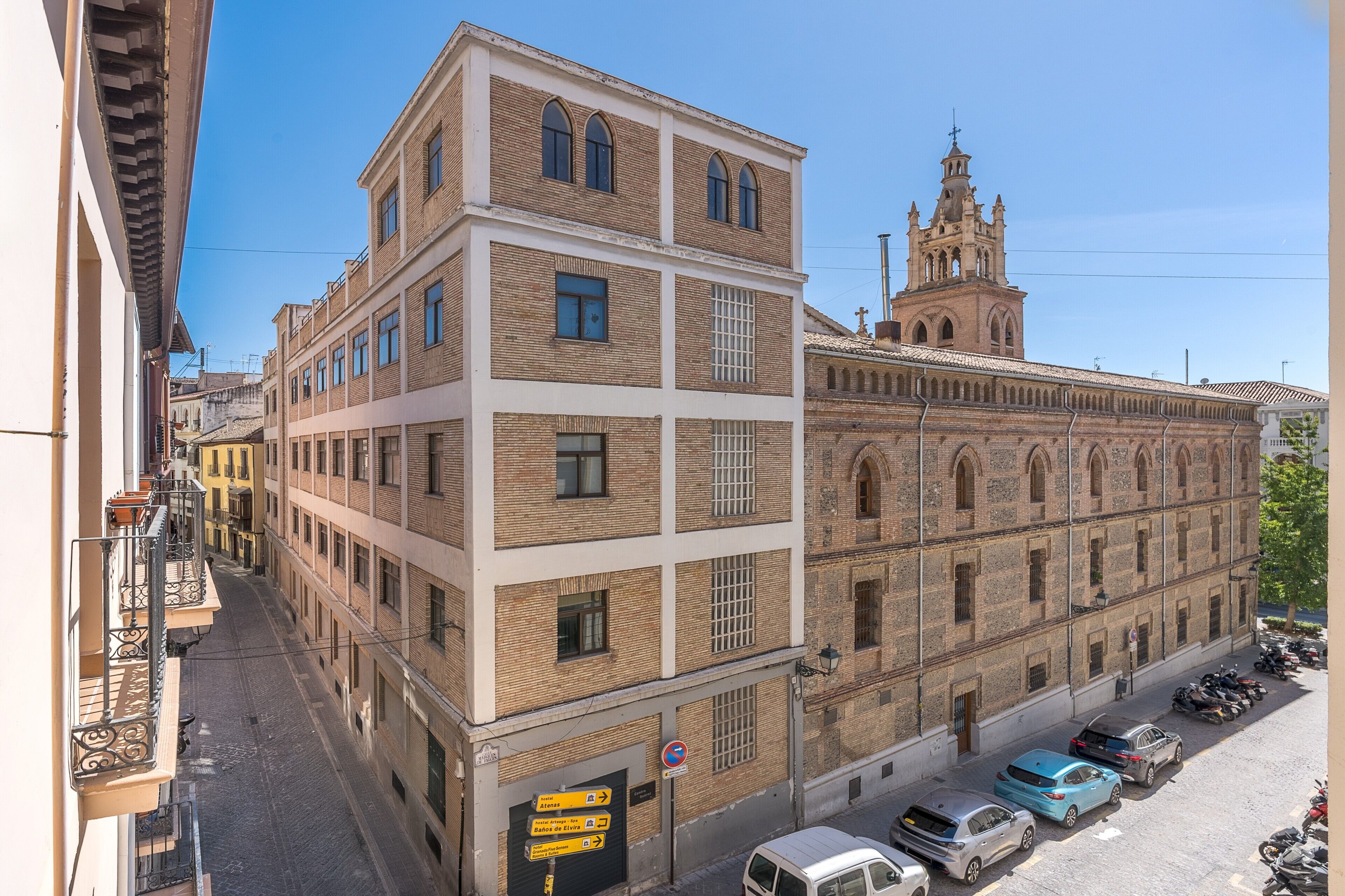 Un edificio de varios pisos con una imponente torre de iglesia, una calle adoquinada y coches estacionados.