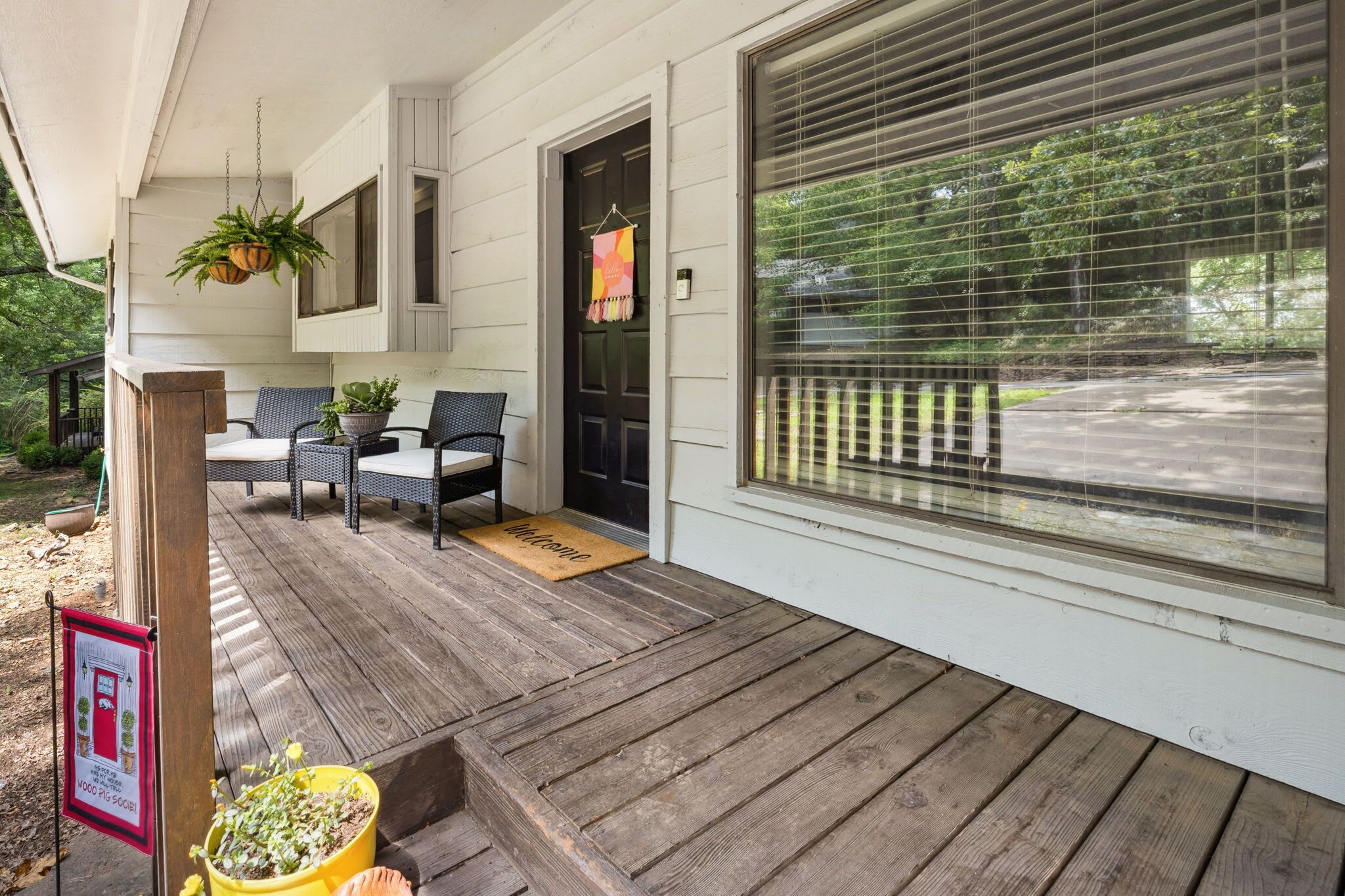 A wooden porch with outdoor furniture, a welcome mat, and potted plants.