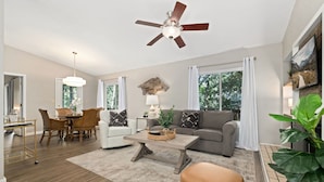 Un salon spacieux doté d’un ventilateur de plafond, d’un canapé gris, d’une table basse et d’un coin repas avec une table et des chaises blanches.