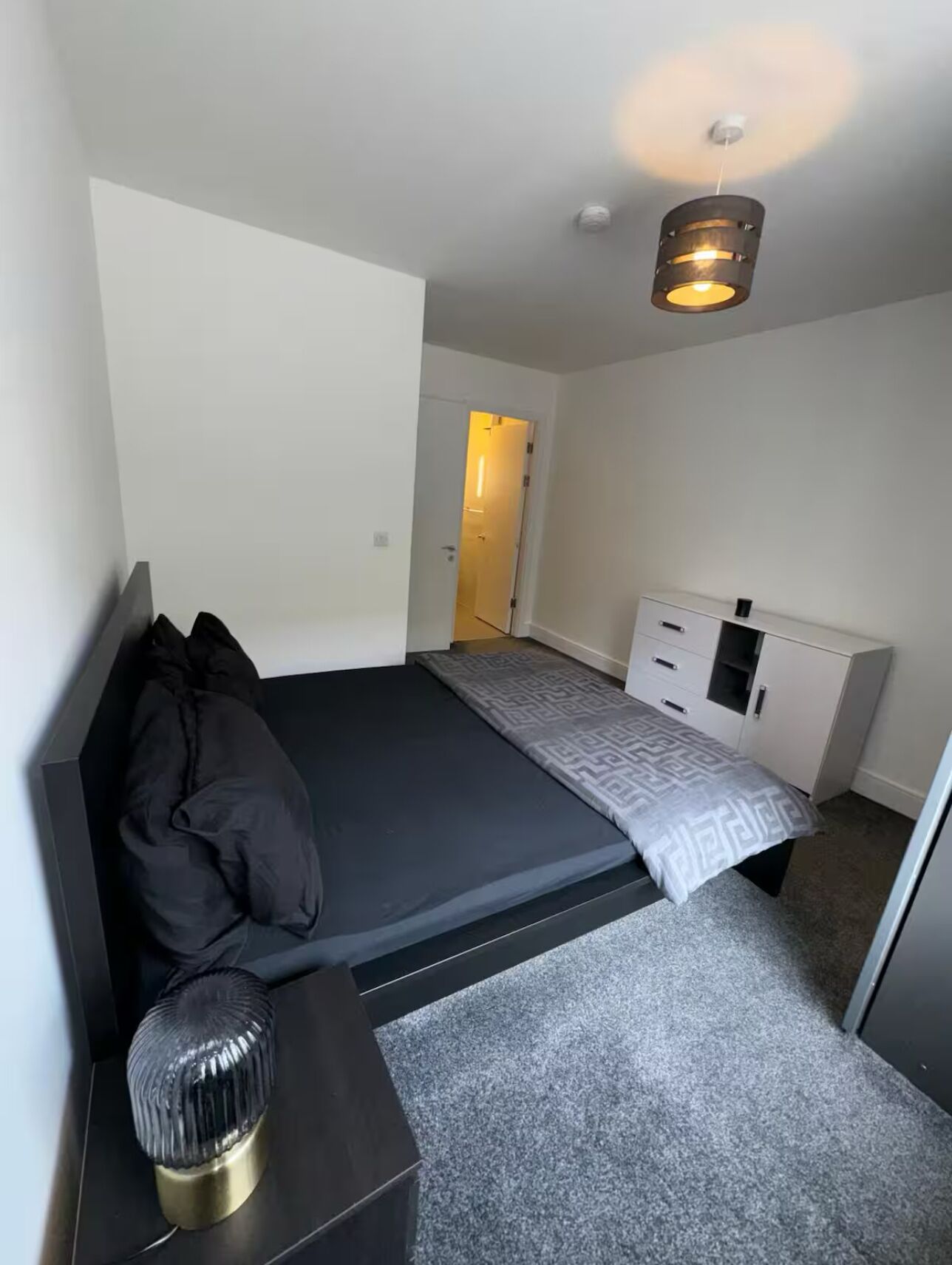 A bedroom with a bed, bedside table, lamp, wardrobe, and a door leading to another room.