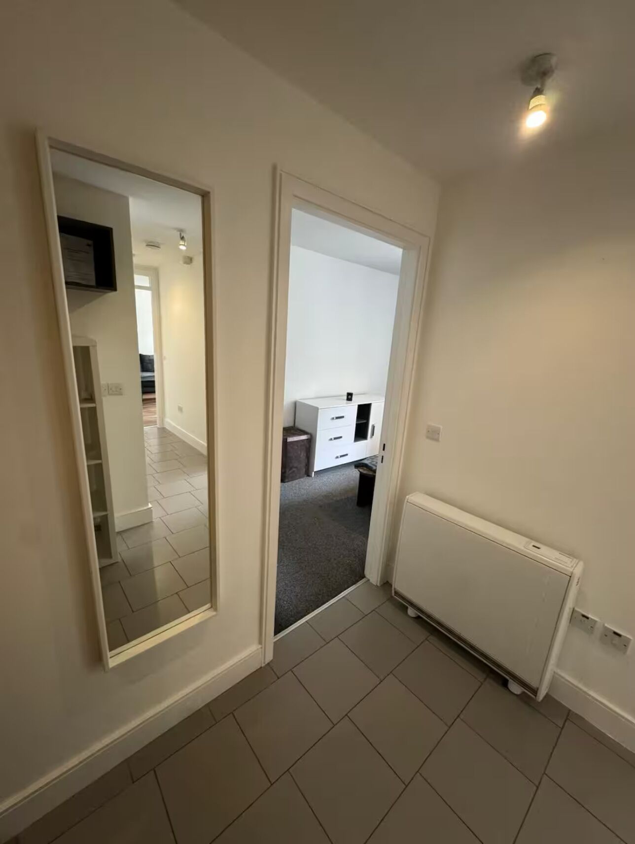 Een gang met een witte radiator, een deur naar een keuken, en een spiegel die de ruimte weerspiegelt.