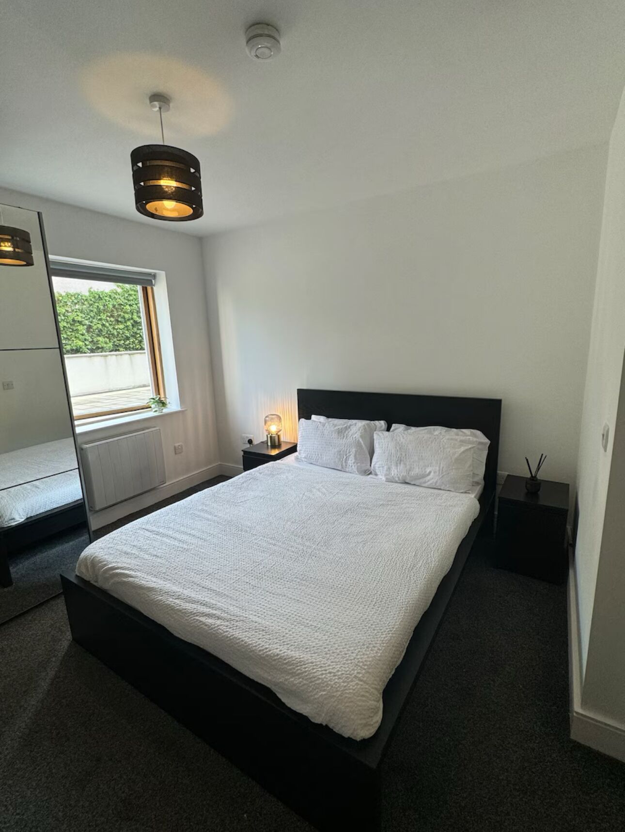 A modern bedroom with a large bed, a bedside table with a lamp, a window with a view, and a ceiling light fixture.