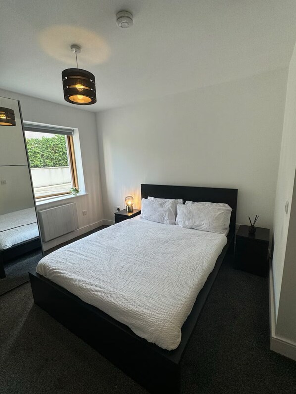 A modern bedroom with a large bed, a bedside table with a lamp, a window with a view, and a ceiling light fixture.