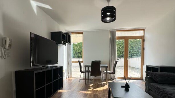 A modern living room with a flat-screen TV, a black entertainment unit, a sofa, a coffee table, and a dining area with chairs and a table.