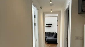 A narrow hallway with white tiled flooring, leading to a room with a gray sofa and a white door.