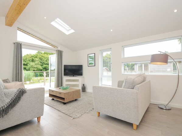 A modern living room with a flat-screen TV, two grey sofas, a wooden coffee table, and large windows with blinds.