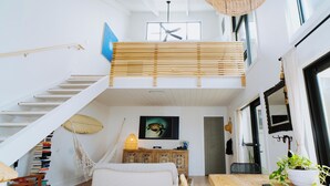 A spacious loft with a high ceiling, a wooden staircase, a large bed, a dining table, and a view of the sky through a window. - Walk to the Beach! Boho-Inspired 2BR Jupiter Loft w Private Outdoor Space (Jupiter)