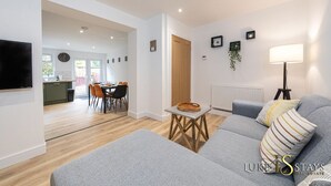 A modern living room with a grey sofa, a dining area with a table and chairs, and a kitchen in the background.