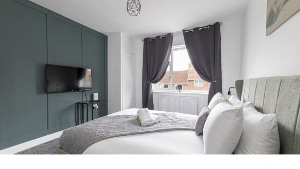 A bedroom with a bed, a flat-screen TV mounted on the wall, a bedside table with a lamp, and a window with curtains.