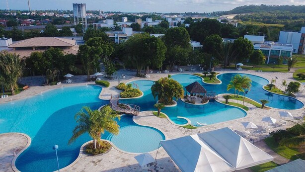 Un gran complejo de piscinas con múltiples carriles, palmeras y zonas sombreadas, rodeado de edificios y follaje.
