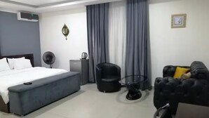 A modern hotel room with a bed, a seating area with a black sofa and armchairs, a small table, and a fan. - Super Powered Mansion  Room 24hr AC & Power (Ikeja)