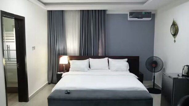 A hotel room with a large bed, a bedside table with a lamp, a standing fan, and an air conditioning unit on the wall. - Super Powered Mansion  Room 24hr AC & Power (Ikeja)