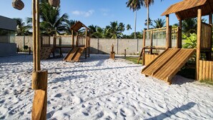 Zona de juegos al aire libre con arena y estructuras de madera, como toboganes y columpios, rodeada de palmeras.
