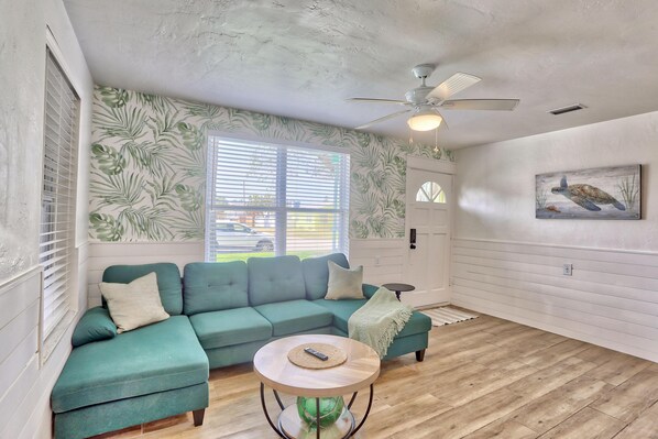 A living room with a teal sectional sofa, a round coffee table with a remote, and a ceiling fan.