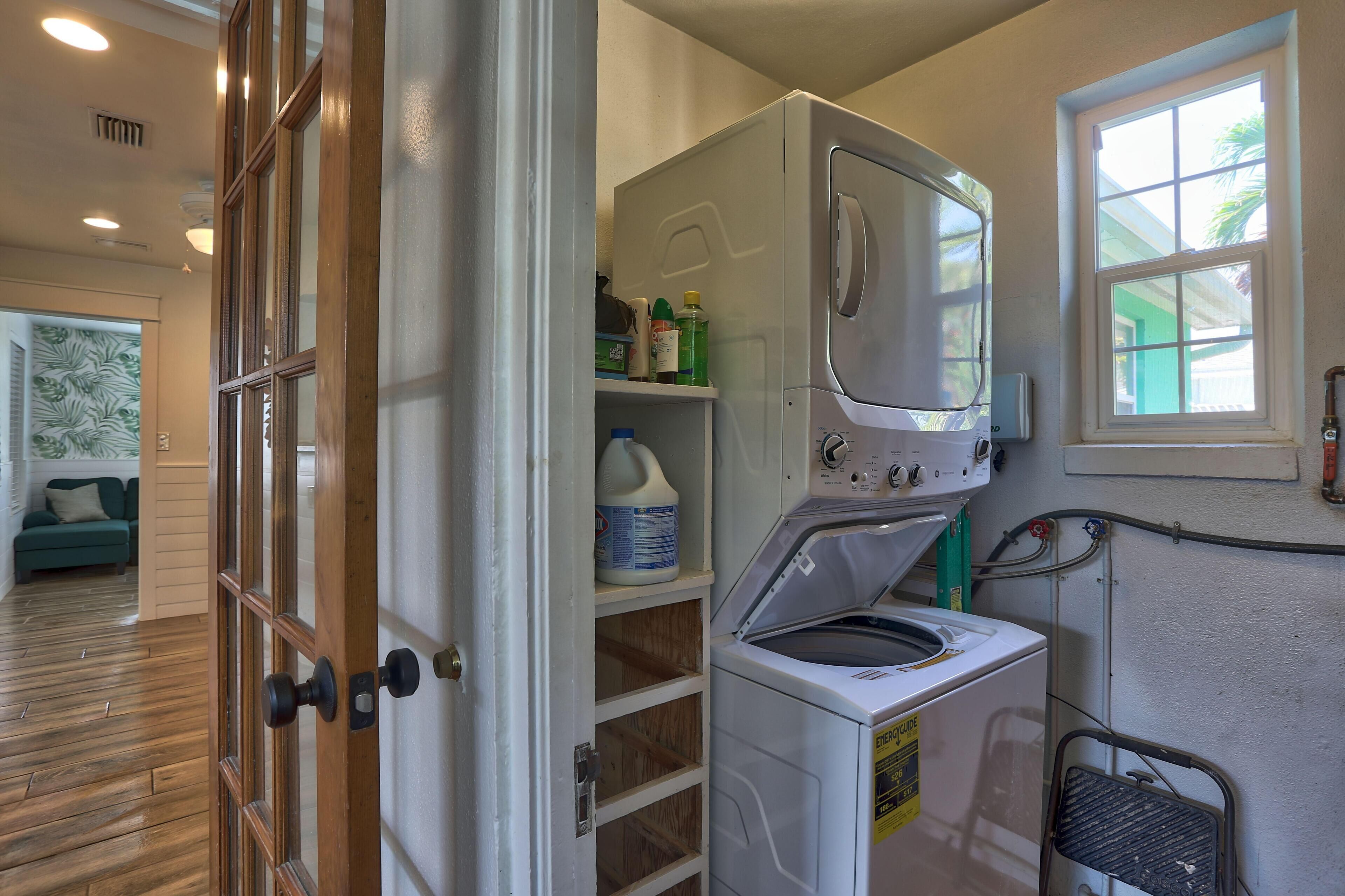 A laundry room with a stacked washer and dryer, shelves with cleaning supplies, and a window with a view of the outside.
