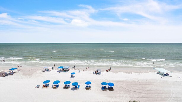 A beach with rows of blue umbrellas and lounge chairs, a sandy shore, and the ocean in the background. - Ram Sea I 405 (North Redington Beach)