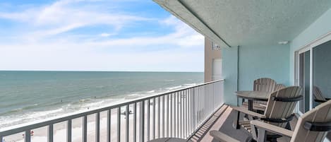 A balcony with a view of the beach and ocean, equipped with tables and chairs.