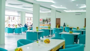 Een ruime eetruimte met teal stoelen en witte tafels, met een modern plafondontwerp en een zichtbare keuken op de achtergrond.