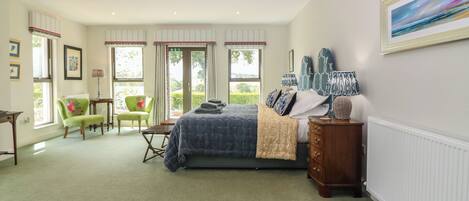Une chambre spacieuse avec un grand lit, une commode, une chaise et un bureau. Elle est dotée de fenêtres avec des rideaux et offre une vue sur la verdure environnante.