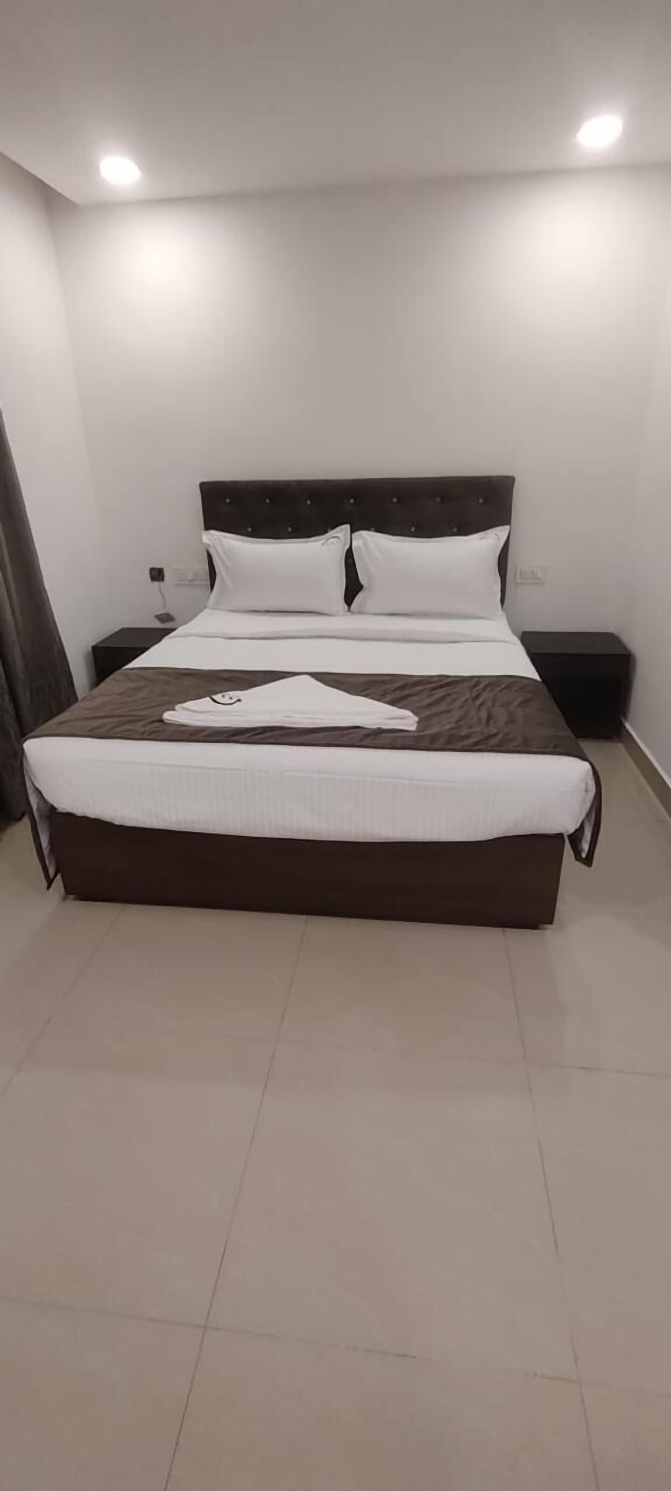 Una cama bien hecha con cabecera oscura y ropa de cama blanca, una mesita de noche con lámpara y un enchufe en la pared.