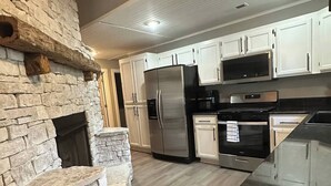 A modern kitchen with white cabinets, a stone fireplace, stainless steel appliances, and a dark countertop. - Fireplace Lodge - 1 mile from Golf Course & 5 mins from the lake! (Isabella)